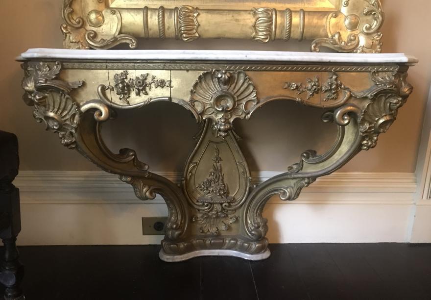 Carved gilt marble-topped table, the top of which forms a half turtle shape.  Lyre-shaped back with two curved supports extending from the base, which rests on a marble piece.  Floral designs on front panel and large flowers decorate the corners and center panel at the top of the table.  