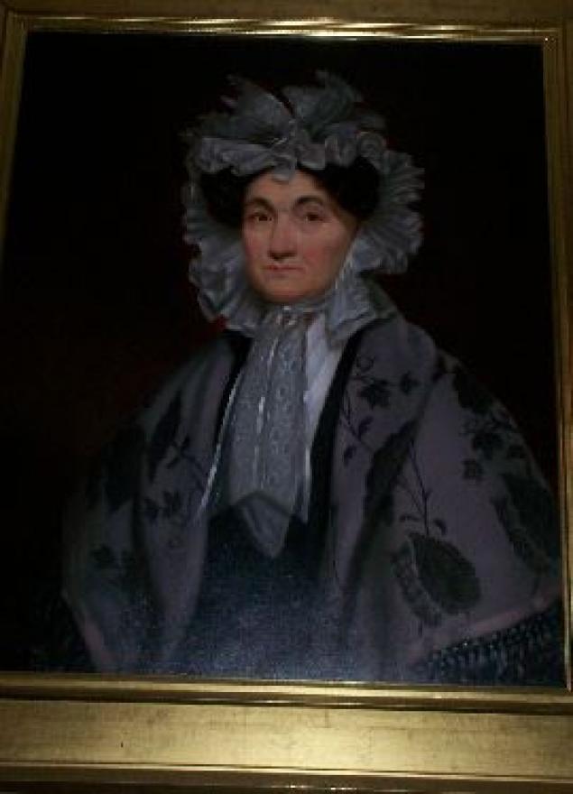 Older woman with brown eyes and dark hair braided up and wearing a light white/grey bonnet.  Draped over her shoulders is a grey fringed shawl with darker crewel-like designs.  Underneath she is wearing a dark garment with a white scarf at her neck.  The background is a solid dark brown. 