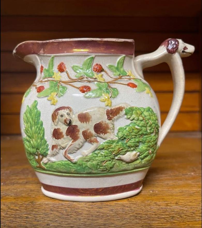 Milk pitcher trimmed in gold lustre.  Majolica-like material with three dogs in relief on sides and dog's head on handle.  Gold lustre band around top and bottom.  Cracking on spout.