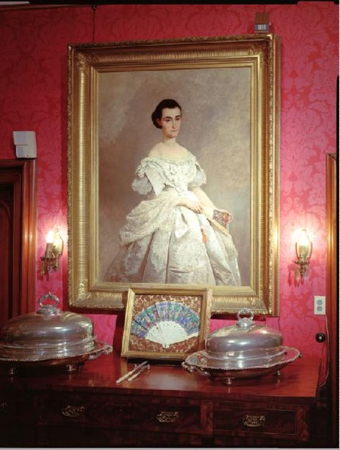 A 3/4 length figure in white, brocaded satin dress holding fan.