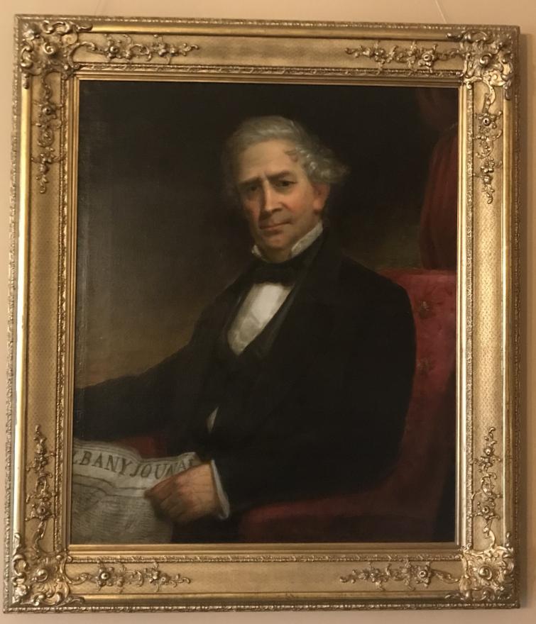 older gentleman with white/grey hair in a suit jacket, starched white shirt, and starched black bow-tie. He sits in a red arm chair in front of a single draped red curtain holding the Albany Journal. He looks at the viewer with a furrowed brow as if he is concerned