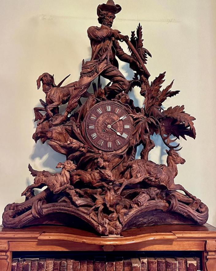 Carved with a scene of a hunter and three dogs chasing a deer around the circular clock base.  The clock has white Roman numerals and white hands. 