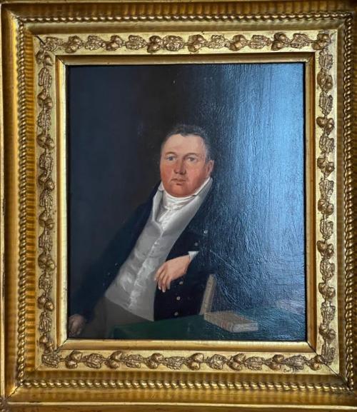 Young man with close-cropped hair wearing a white shirt with a cravat, a grey waistcoat, and a dark blue jacket. His cheeks are a ruddy red as if the wind has chapped his cheeks. He leans back in a chair with his left arm draped over the back of a wooden chair. He sits behind a table with a green table cloth and a beige leather book on top of the table.
