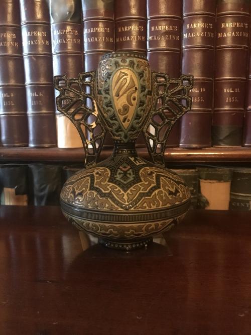 Ceramic urn with black background with ochre, yellow, blue, green and pink colors in an arabesque style. The top of the urn is bulbous with a bell bottom and two handles. 