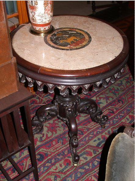 Marble top has a scene with a horse and a palm tree in yellow, red and gray in a circular center with the inscription "Carthago Delenda Est."  Top of table has a Gothic fret-carved border with turned pendants, on a leaf-carved shaft.  Quadrupedal base, each leg with double-scroll design.